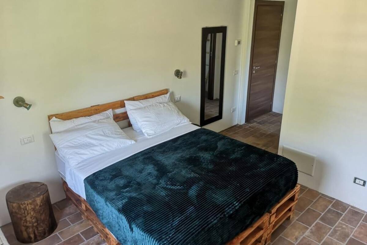 A bedroom with a wooden bed, white bedding, a dark green blanket, a mirror, and a wooden stool.
