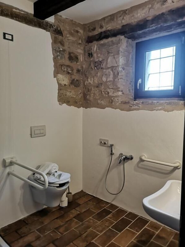 A bathroom with a stone wall, a window, a toilet with a grab bar, a shower, and a sink. - Casa Giogano (San Leo)