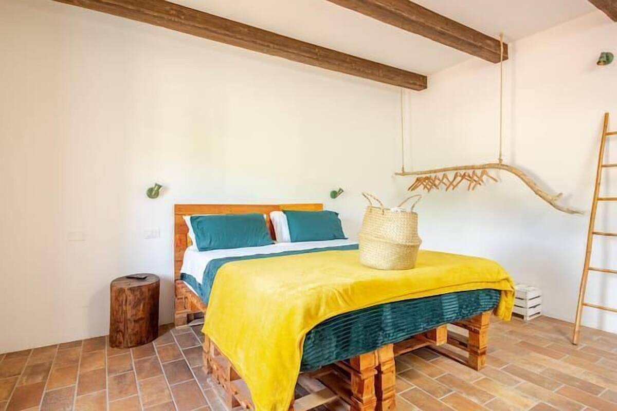 A room with a bed, a wooden bench, a woven basket, and a ladder.