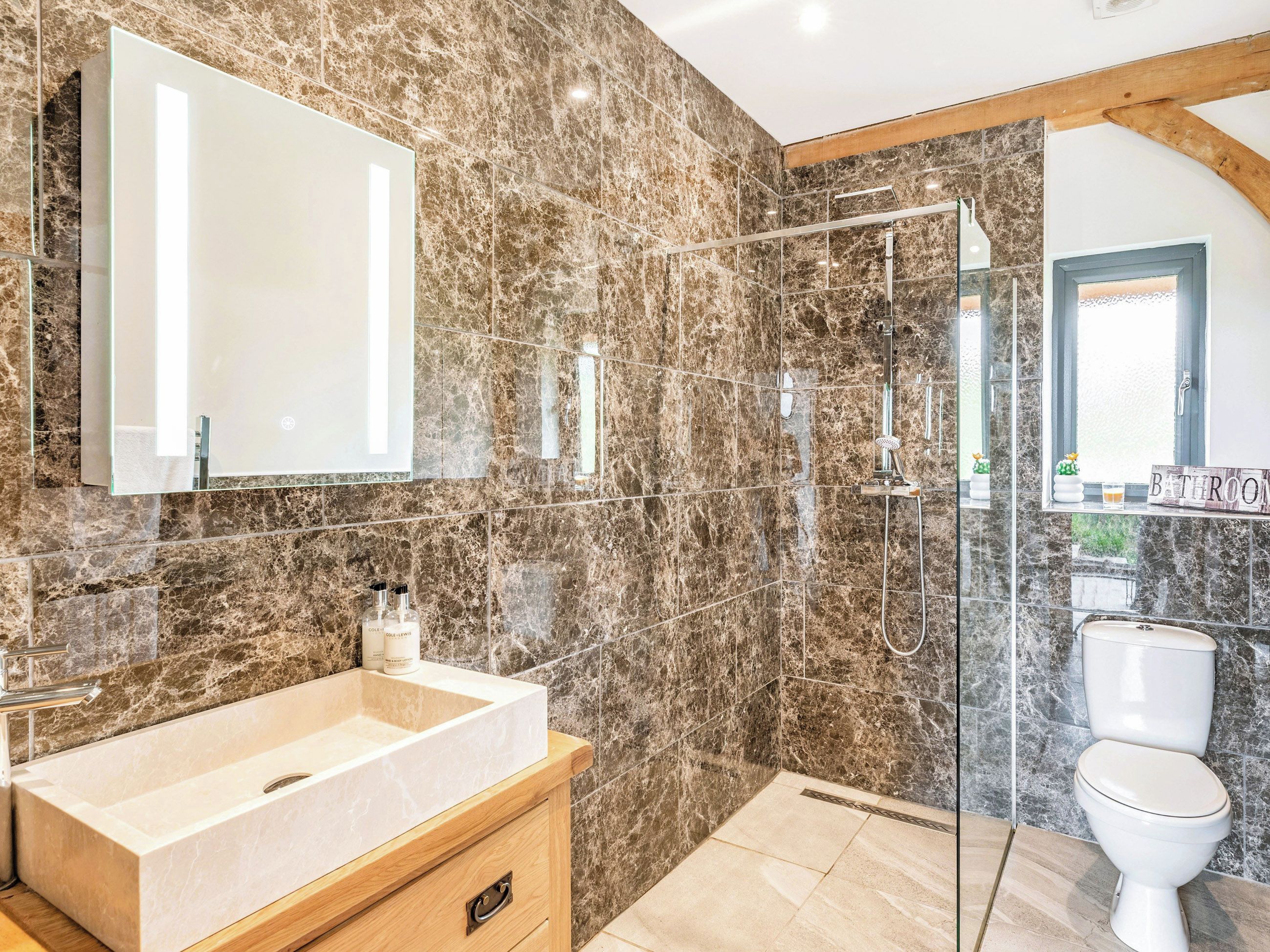 A bathroom with a marble wall, a large mirror, a sink, a shower, and a toilet.