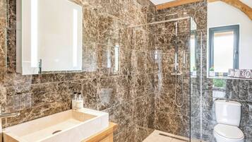 A bathroom with a marble wall, a large mirror, a sink, a shower, and a toilet.