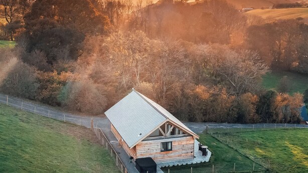 Een houten hut met een schuin dak, omgeven door een omheind erf en weelderige begroeiing.