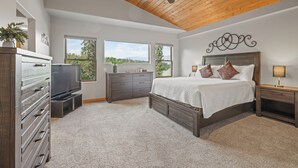 A bedroom with a wooden ceiling, a bed with white bedding, a dresser, a TV, and two windows offering a view of trees.