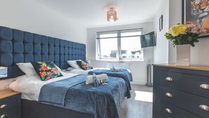 Moderni hotellihuone, jossa on koristeltu päätyseinä, suuri ikkuna, josta on näkymä, taulutelevisio ja lipasto laatikoineen.
