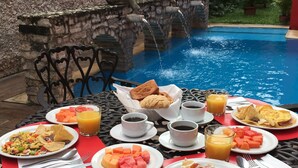 Une table à manger dressée avec des aliments pour le petit-déjeuner, notamment des œufs, des toasts et des fruits, le tout disposé devant un décor de piscine agrémenté d’un mur en pierre et de plantes décoratives.