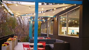 An outdoor seating area with tables and chairs, a pergola with string lights, and a view of a restaurant in the background. - Tru By Hilton Chambersburg, PA (Chambersburg)
