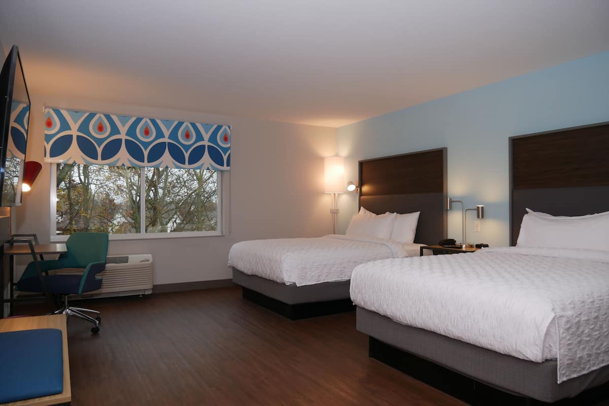 A hotel room with two beds, a desk, a chair, and a window with a view of trees.