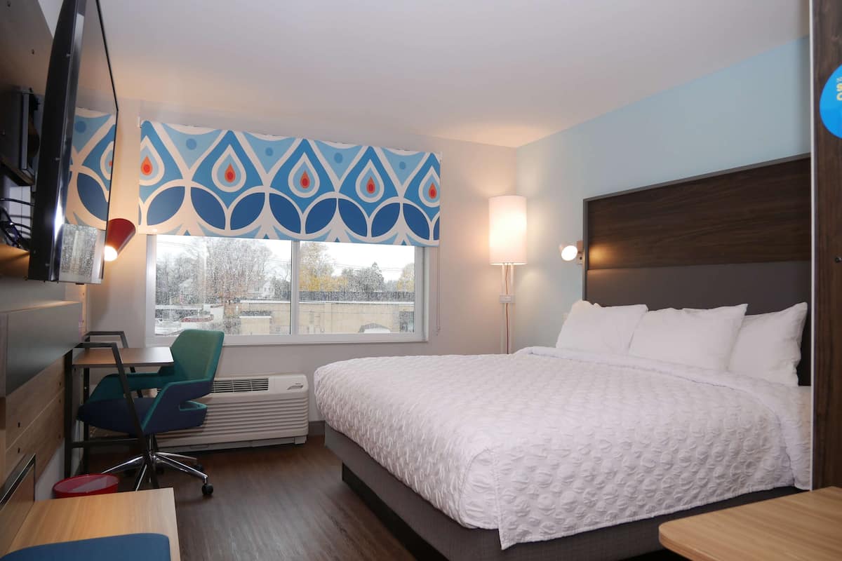 A hotel room with a large bed, a desk with a chair, a window with a view of the city, and a wall-mounted TV.