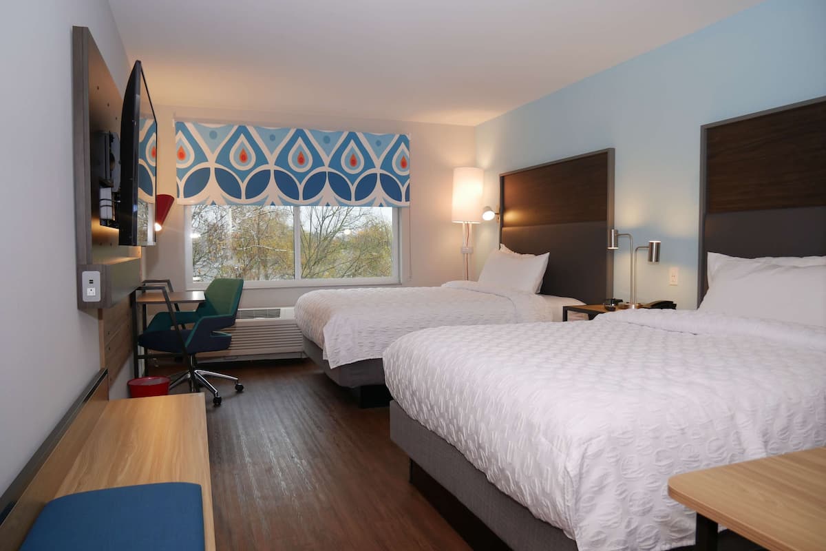 A hotel room with two beds, a desk, a chair, a TV, and a window with patterned curtains.