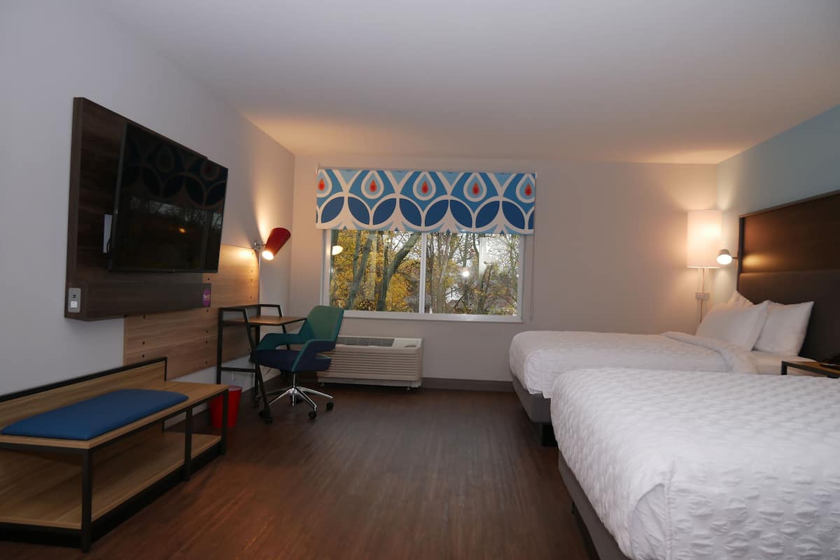 A modern hotel room with a bed, a desk, a chair, a TV, and a window with a view of trees.