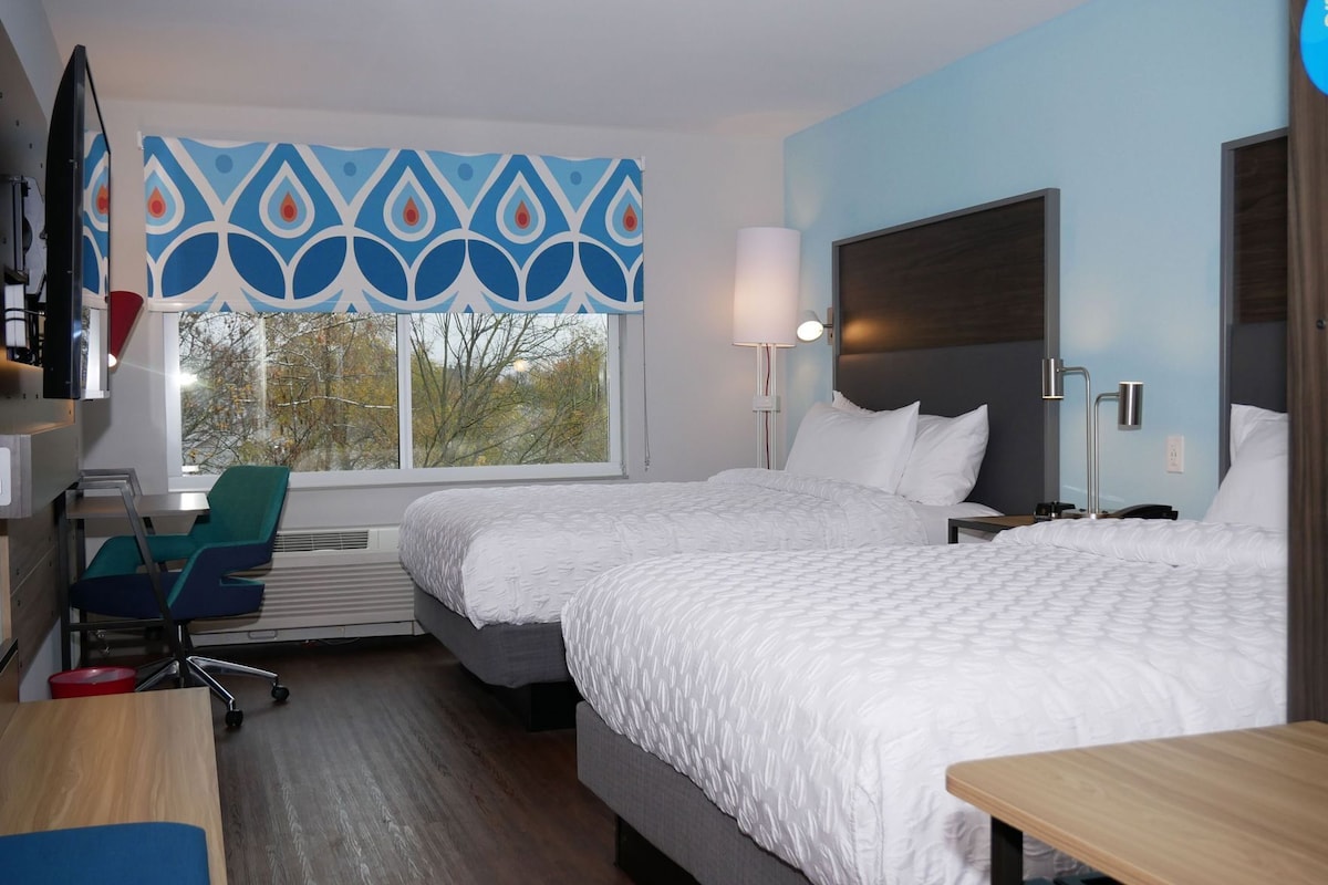 A hotel room with two beds, a desk, a chair, a window with a view, and a decorative wall panel.