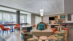 A modern hotel lobby with seating areas, a central sofa with a decorative pillow, and a bar area with shelves and a television. - Home2 Suites by Hilton Panama City Beach, FL (Panama City Beach)