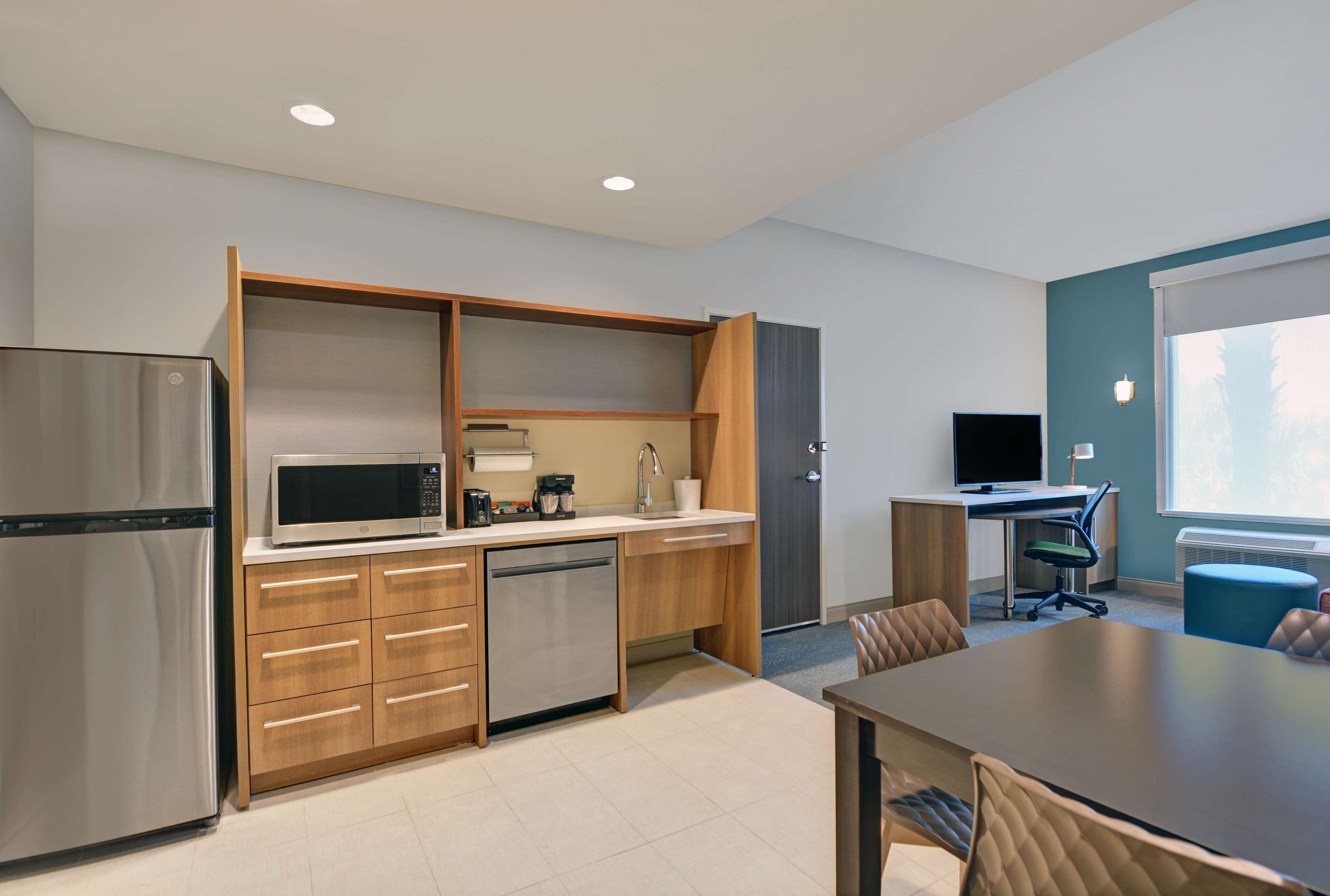 A modern kitchen with a built-in microwave, refrigerator, and dishwasher. There is a dining area with a table and chairs, and a small office space with a desk and computer.