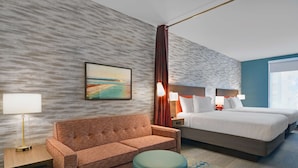 A hotel room with a sofa, a bed, a nightstand, a lamp, and a painting on the wall. - Home2 Suites by Hilton Panama City Beach, FL (Panama City Beach)