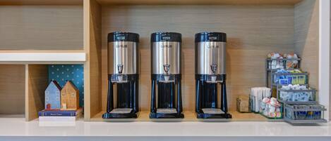 Three coffee makers on a counter, a shelf with a house model and books, and a storage area with mugs and supplies.