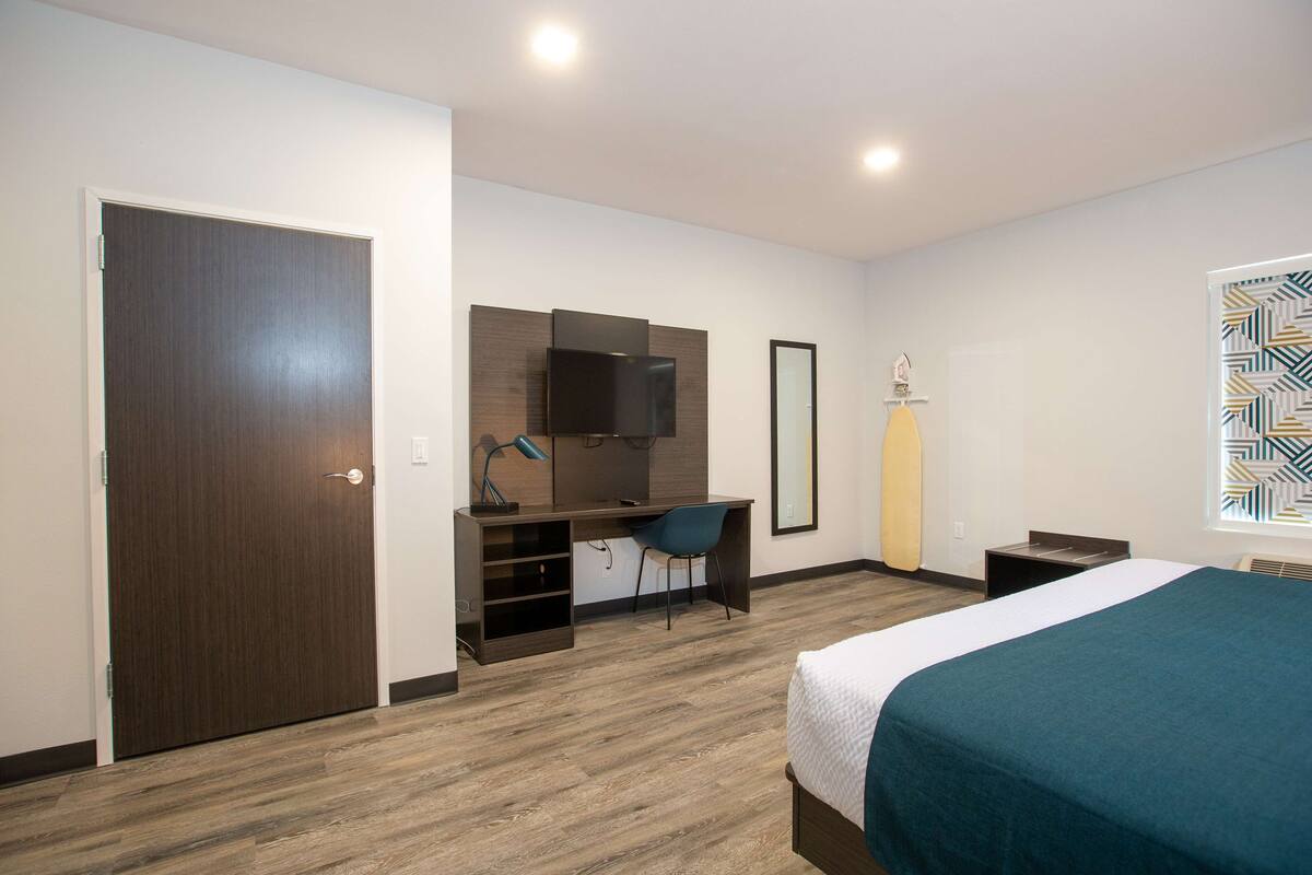 A modern hotel room with a wooden floor, a bed with a teal bedspread, a desk with a chair, a flat-screen TV, and a large window with geometric blinds.