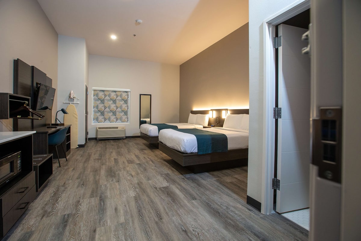 A hotel room with a kitchenette, a bed with a headboard, a desk with a chair, and a large mirror.
