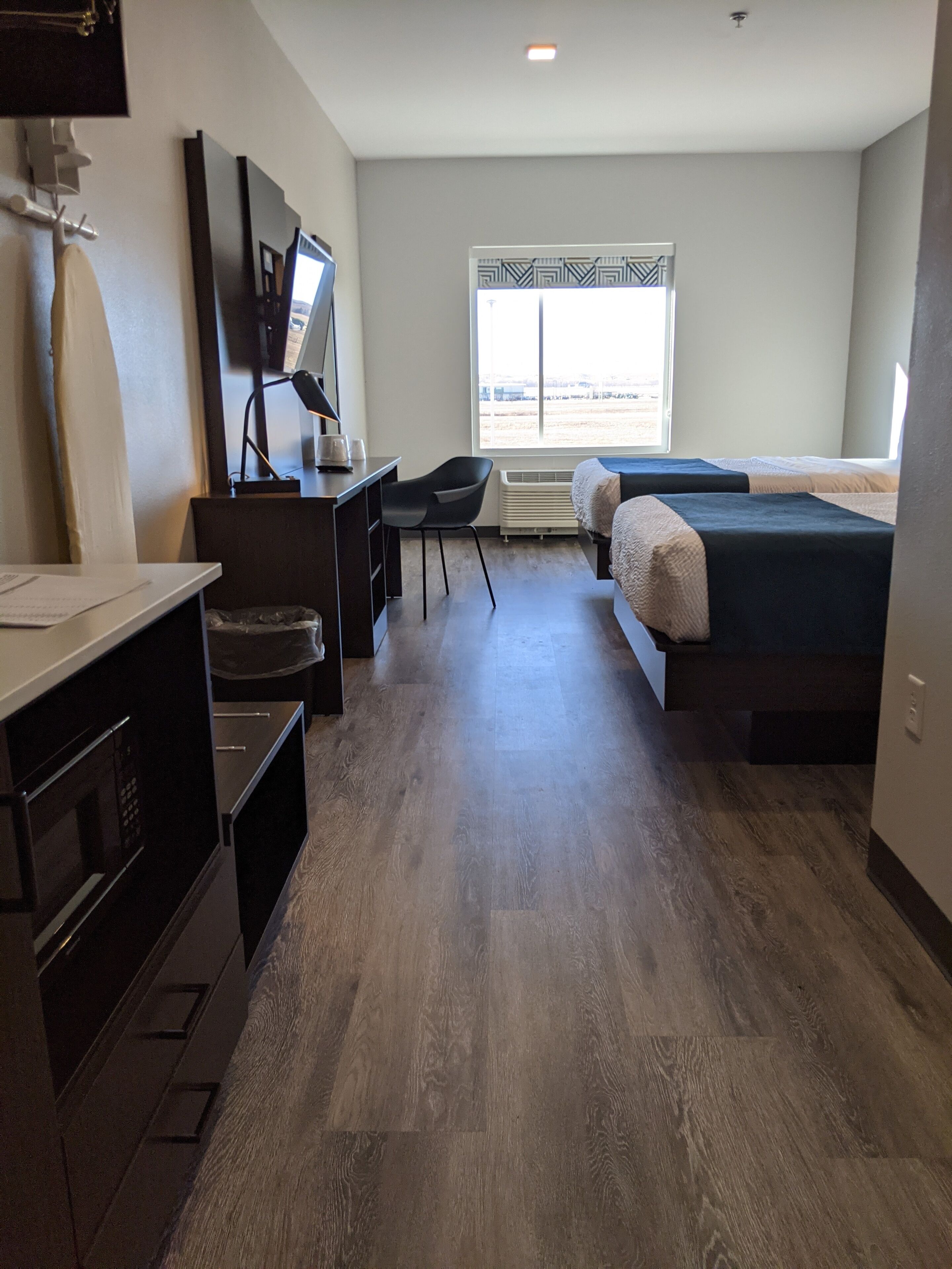 A modern hotel room with a wooden floor, a desk with a chair, a bed, a TV, and a window with a view of the outside.