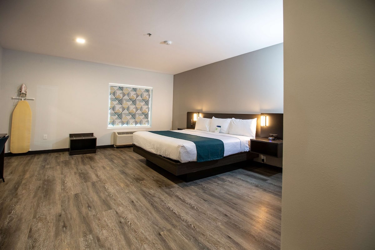 A modern hotel room with a large bed, a window with patterned curtains, a wooden floor, and a small side table.