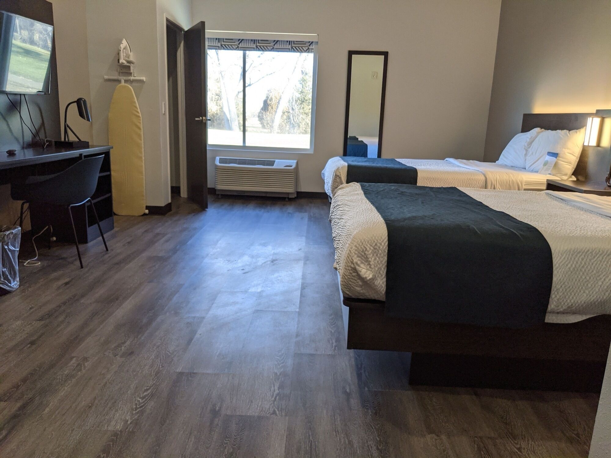 A hotel room with a desk, two beds, a TV, an ironing board, and a window with a view of trees.