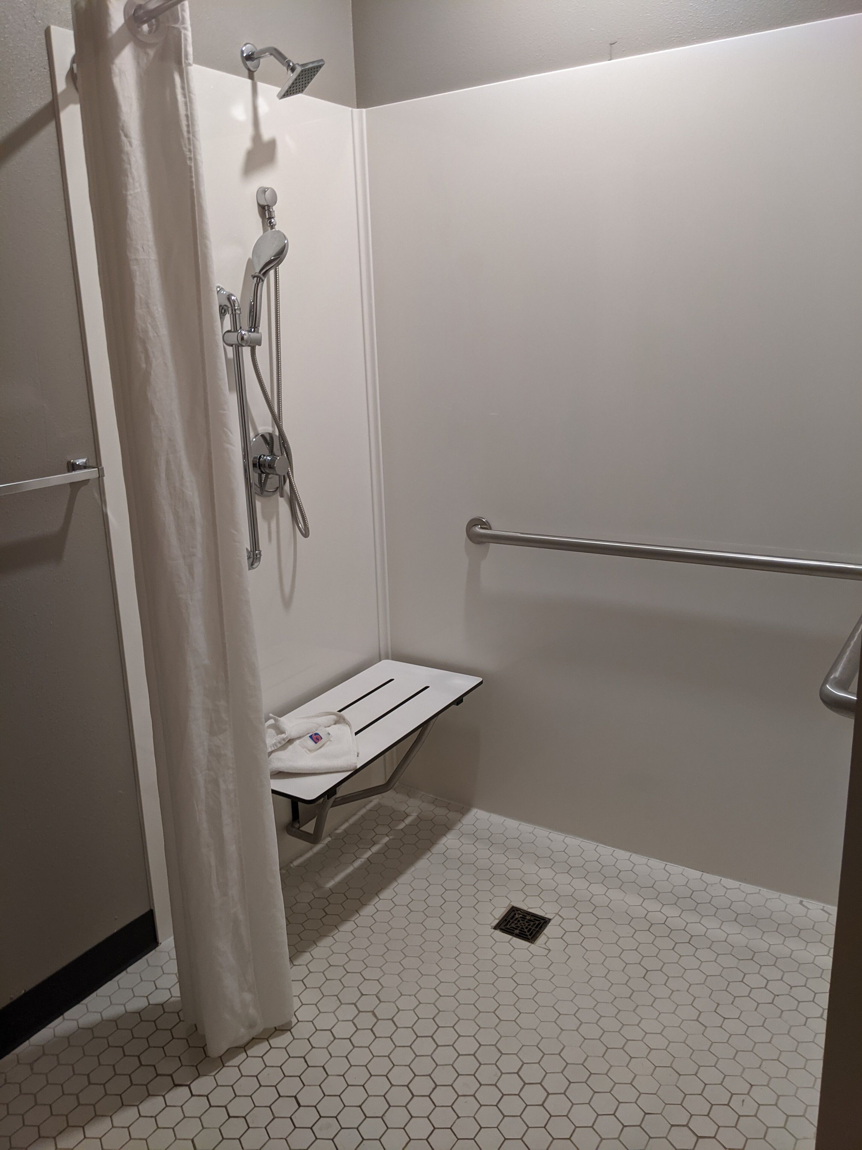 Un petit espace de douche, de couleur blanche, dont le sol est recouvert de carreaux hexagonaux, et qui est équipé d’une barre d’appui, d’un pommeau de douche et d’un porte-serviettes.