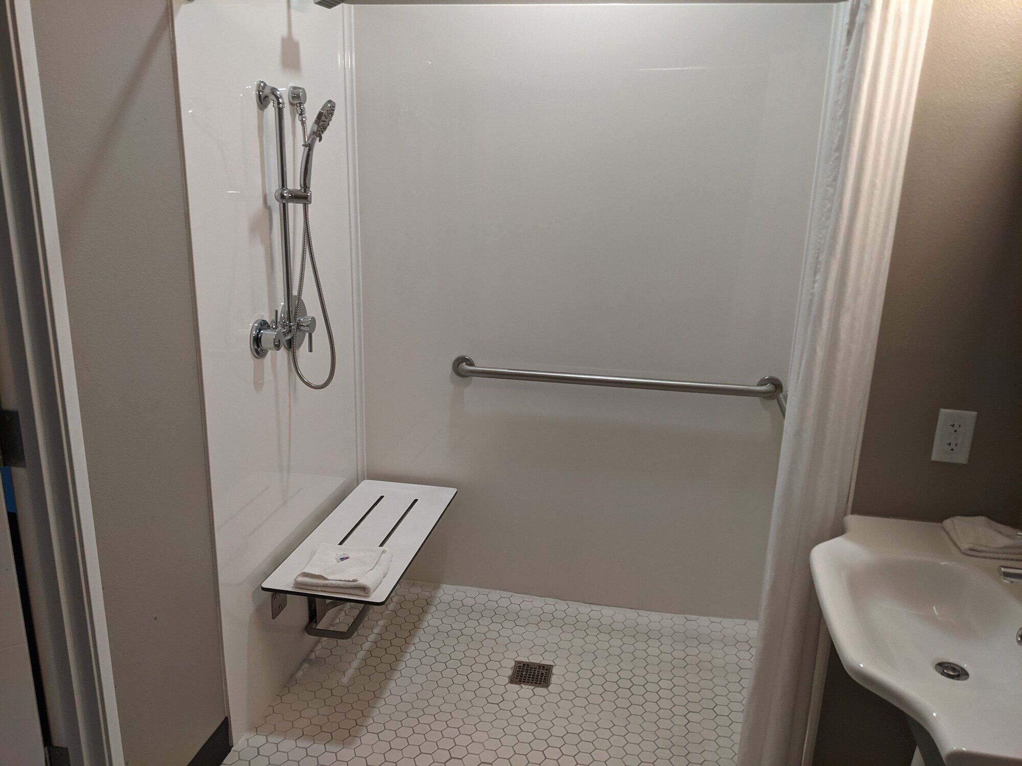 A small, white, walk-in shower with a grab bar, a white sink, and a towel rack.