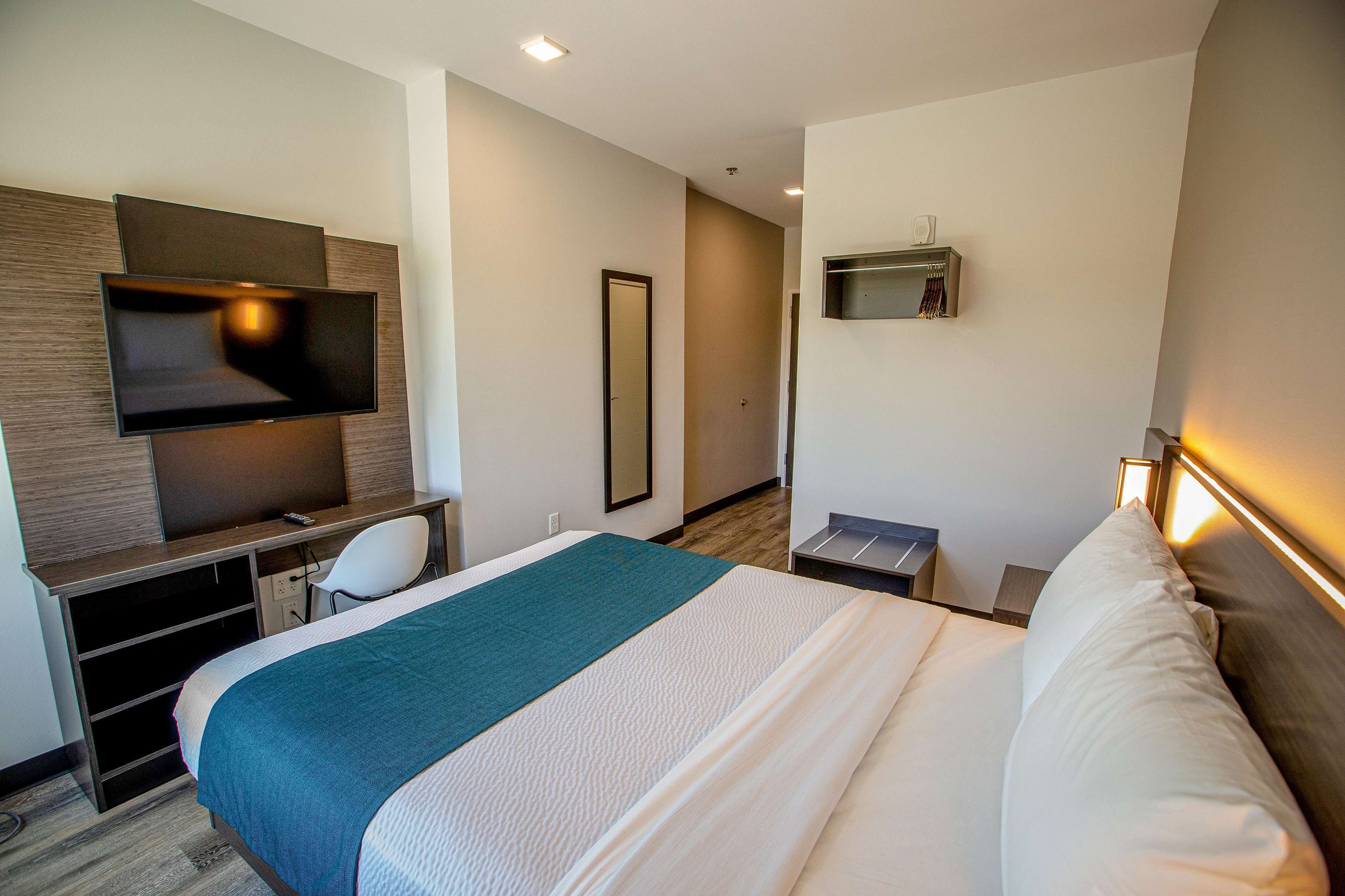 A hotel room with a bed, a desk with a TV, a mirror, and a bedside table.