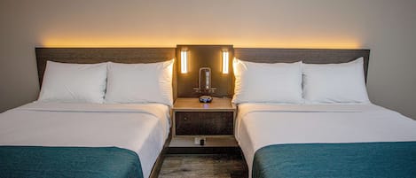 Habitación de hotel con dos camas individuales, cada una con ropa blanca y almohadones color aguamarina. Hay una mesita de noche de madera entre las camas con una pequeña mesa y un reloj.