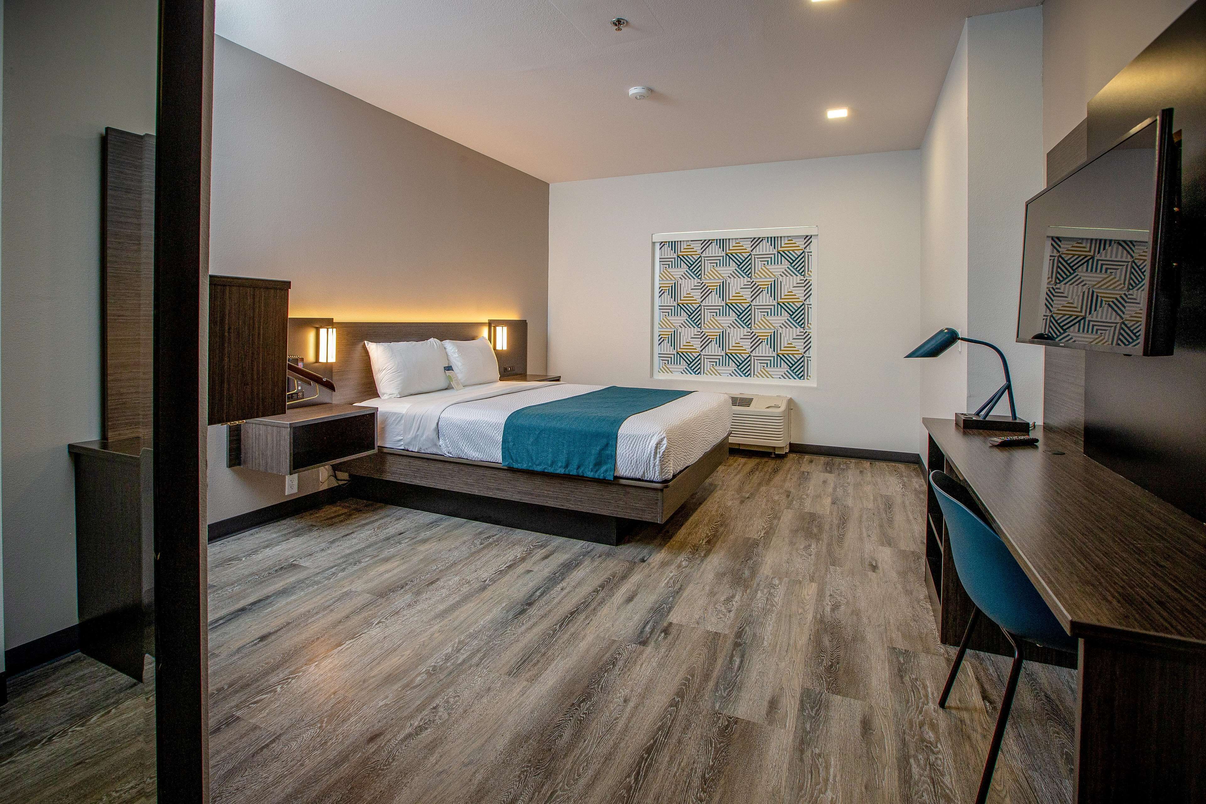 A modern hotel room with a bed, bedside table, desk, chair, and a large mirror.