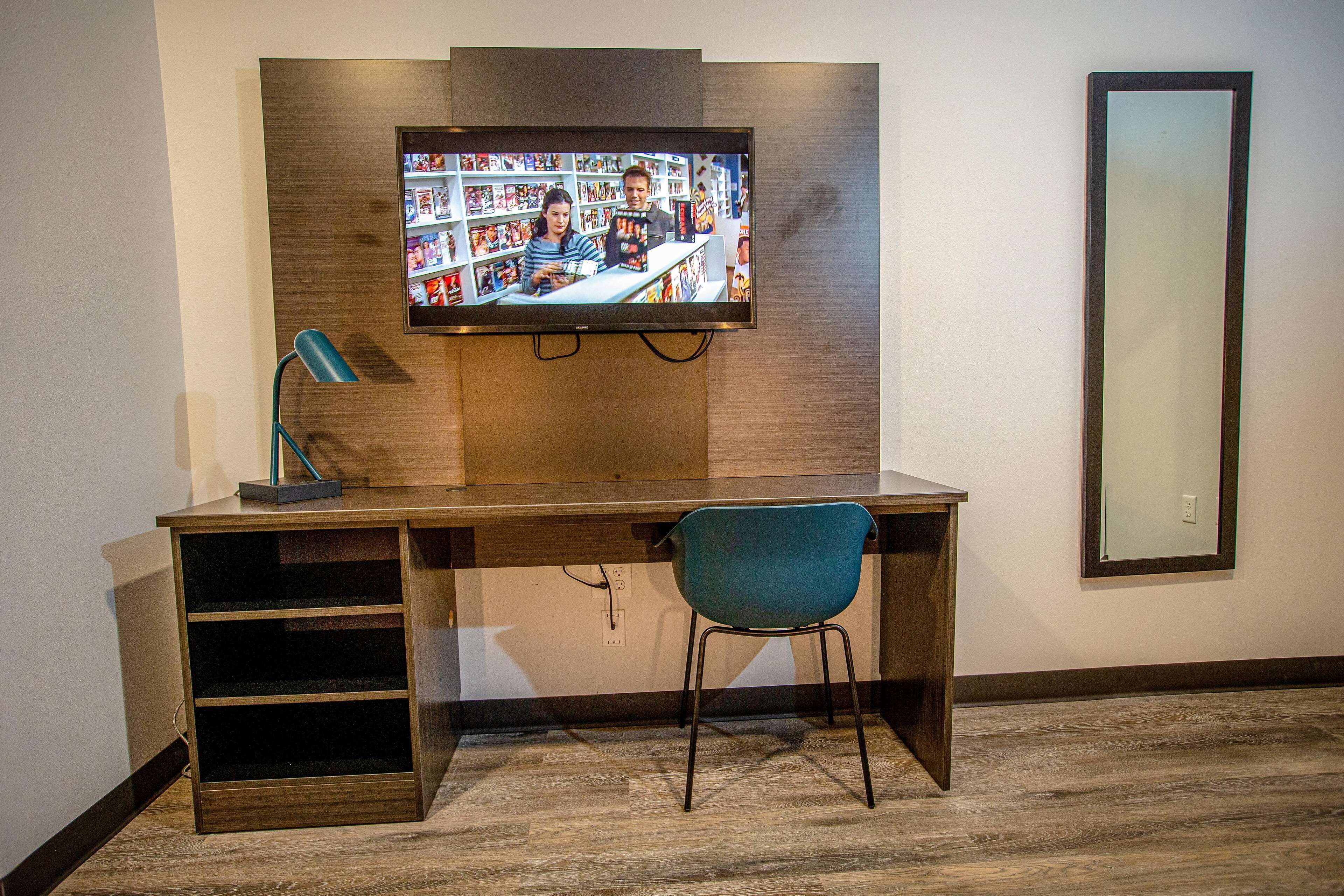 A modern room with a wooden desk, a blue chair, a television mounted on the wall, and a large mirror.
