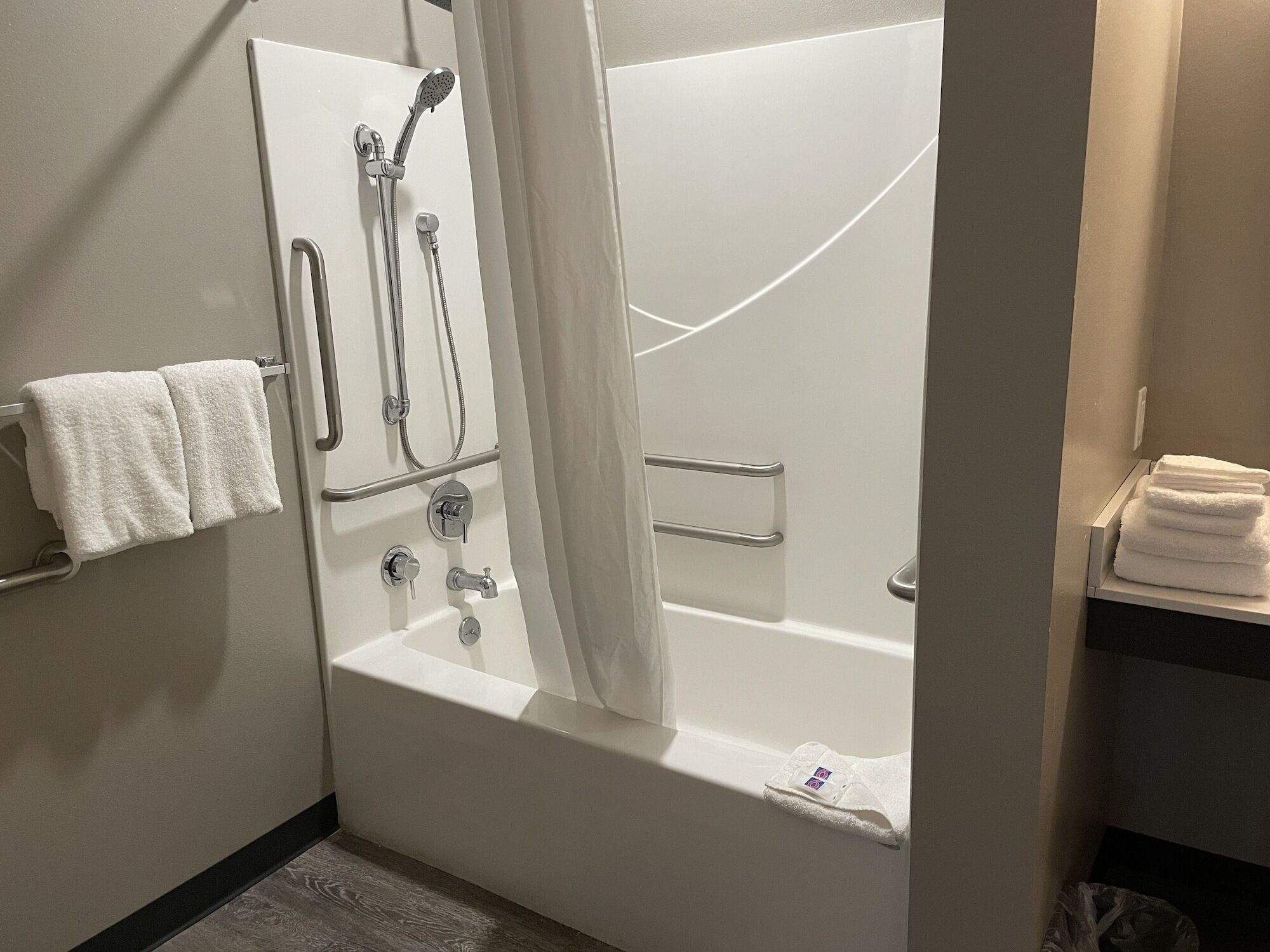 Une salle de bain moderne équipée d’une baignoire, d’une douche et de barres d’appui. On y trouve des serviettes sur un porte-serviettes et une étagère.