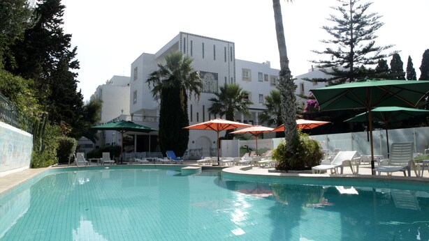 A swimming pool with lounge chairs and umbrellas, surrounded by palm trees and a building with a tiled facade. - Hotel Residence Mahmoud (Hammamet)