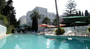 A swimming pool with lounge chairs and umbrellas, surrounded by palm trees and a building with a tiled facade. - Hotel Residence Mahmoud (Hammamet)