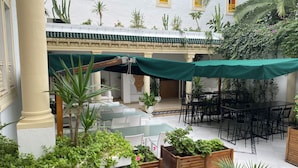 An outdoor patio area with tables and chairs, surrounded by greenery and a white building with yellow window frames. - Hotel Residence Mahmoud (Hammamet)