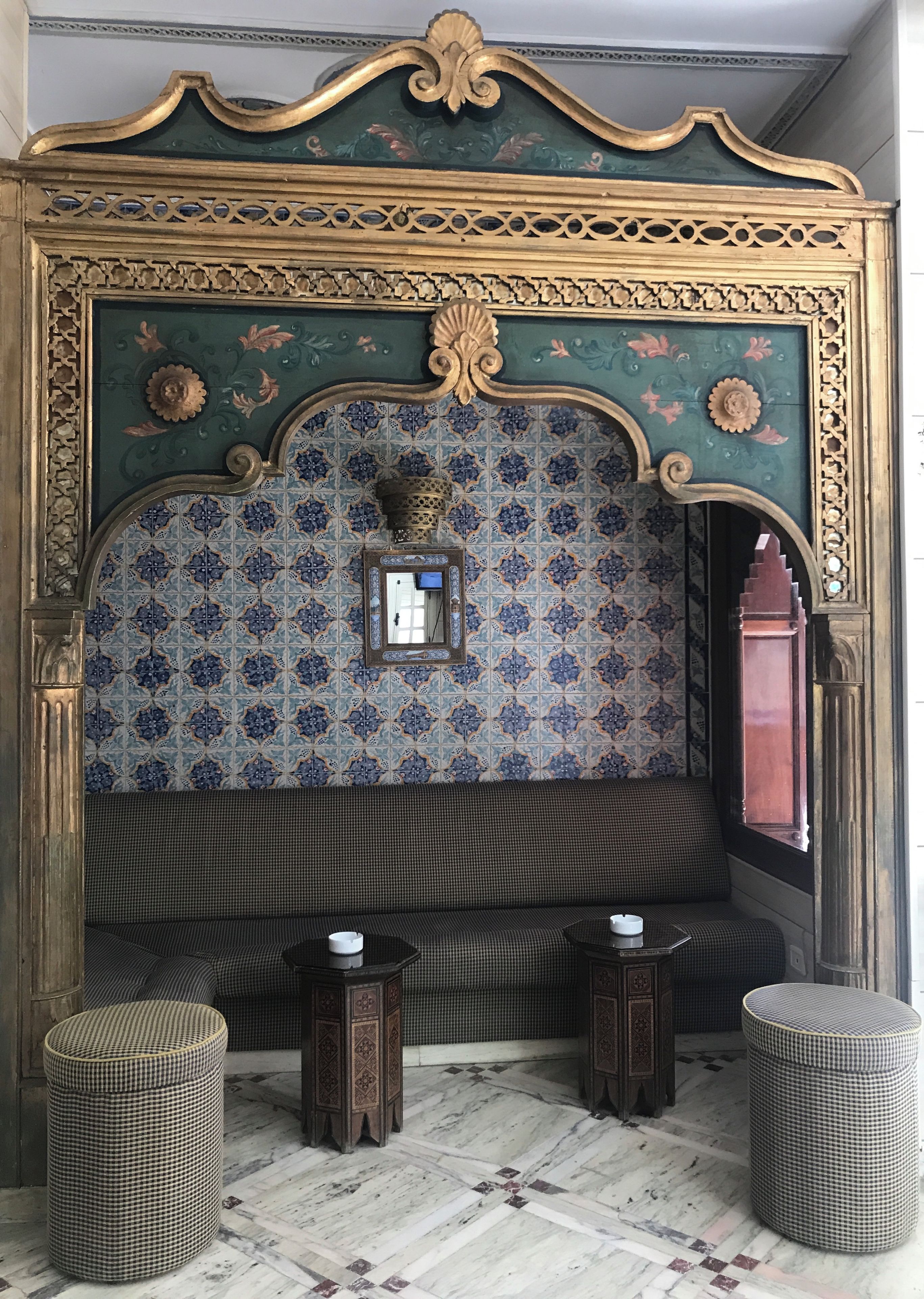 A room with a patterned wall, a cushioned bench, two small tables, and a decorative mirror.