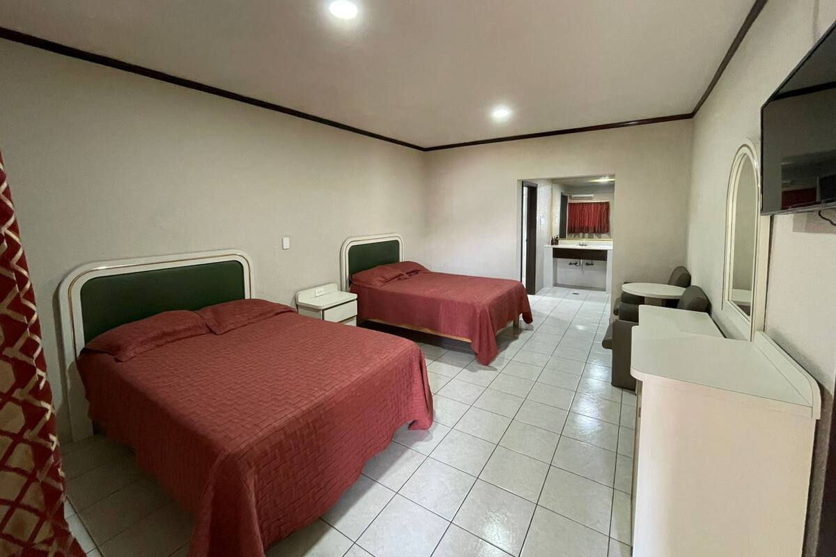 Habitación de hotel con dos camas, un escritorio pequeño, una tocador con espejo y un baño visible a través de una puerta abierta.