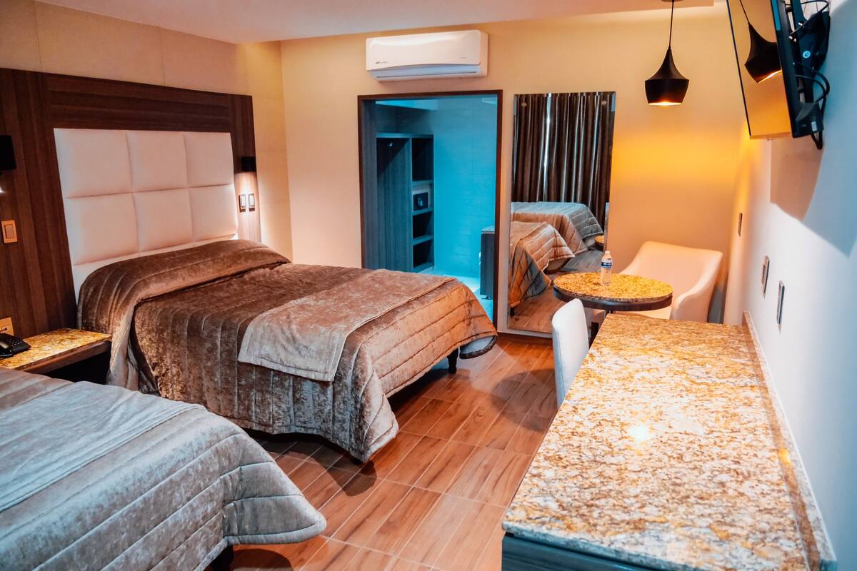 Una habitación de hotel con una cama grande, otra más pequeña, un escritorio, una silla, una mesita y un espejo.