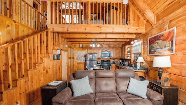 A cozy cabin interior with a wooden staircase, a dining area, and a living room with a brown sofa and two pillows.