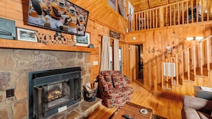 A wooden cabin interior with a stone fireplace, a television mounted above it, and a cozy seating area with a patterned ottoman.
