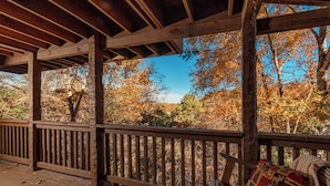 A wooden deck with a railing, a bench, and a view of trees with autumn foliage.