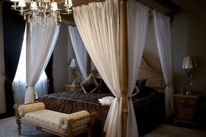 A four-poster canopy bed with a dark patterned bedspread, a wooden nightstand with a lamp, and a chair with a patterned cushion.