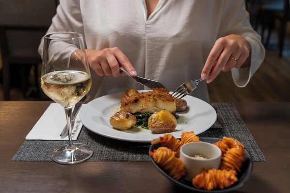 A person dining at a table with a plate of food, a glass of white wine, and a bowl of croissants.
