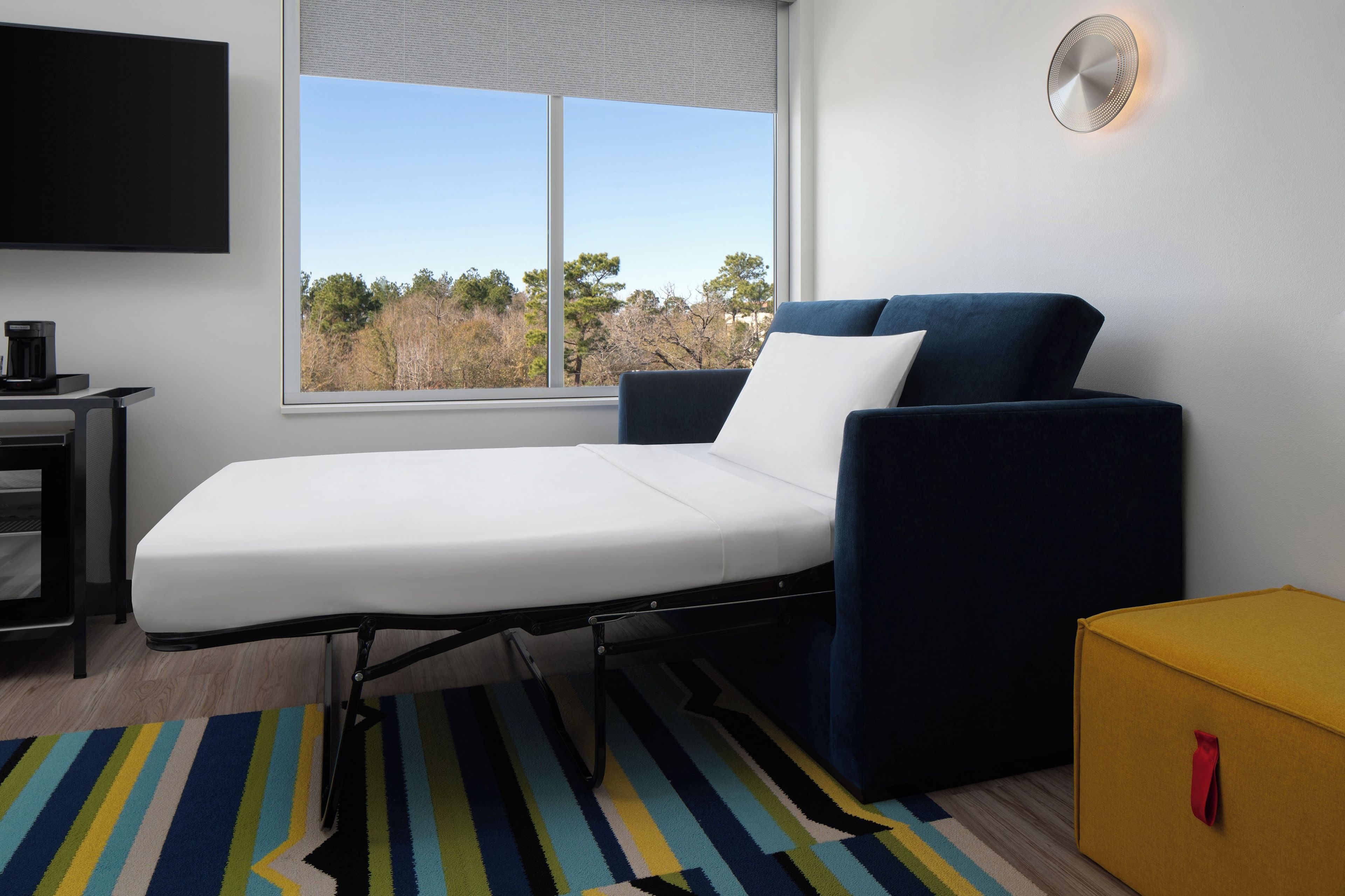 Un lit simple avec un matelas blanc, une tête de lit bleue et un oreiller blanc. On y trouve une petite table sur laquelle se trouve une machine à café, un tapis rayé aux couleurs vives sur le sol et une boîte jaune avec une anse rouge.