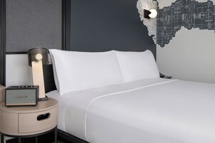 Una habitación de hotel moderna con una cama, una mesita de noche con un altavoz y una pared decorada con un diseño urbano.