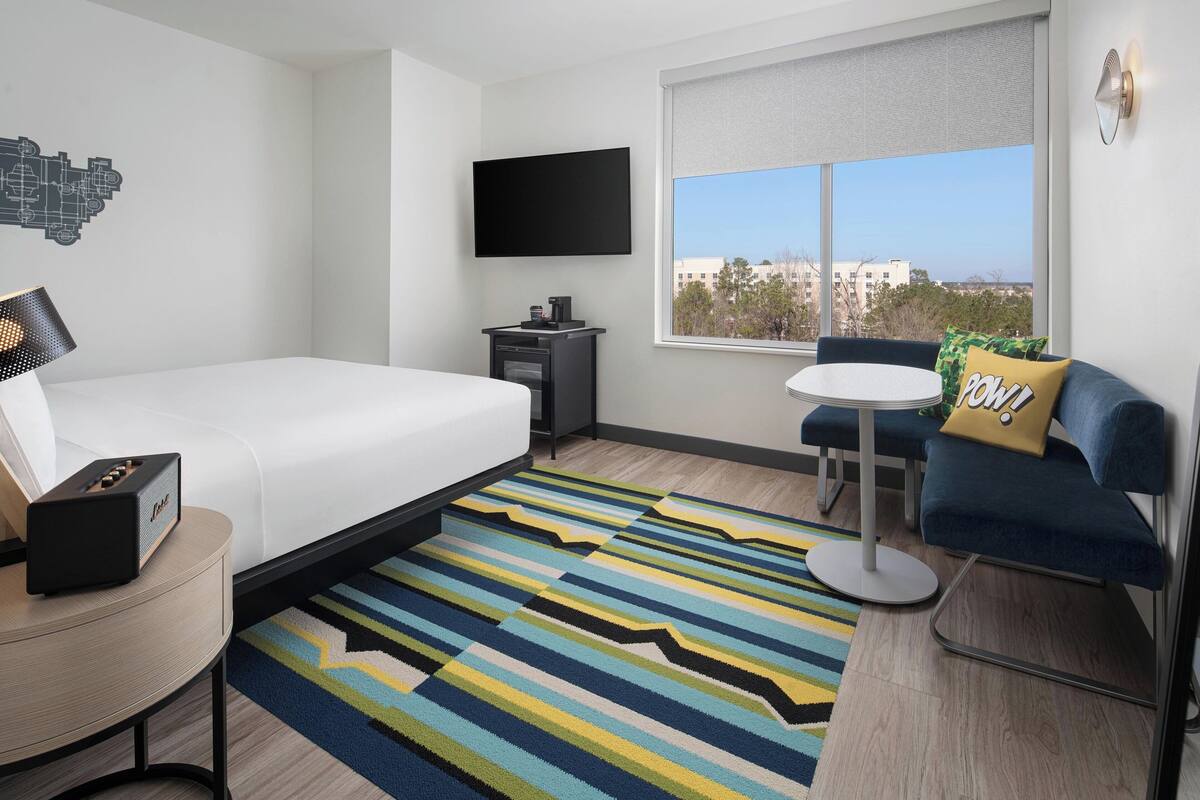 A hotel room with a bed, a desk, a TV, a coffee machine, a window with a view, and a blue sofa with a pillow.