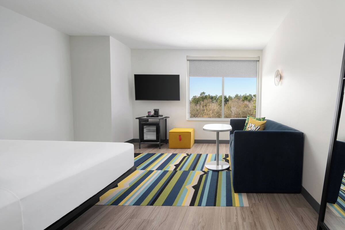 A hotel room with a bed, a sofa, a small table, a TV, and a window with a view of trees.