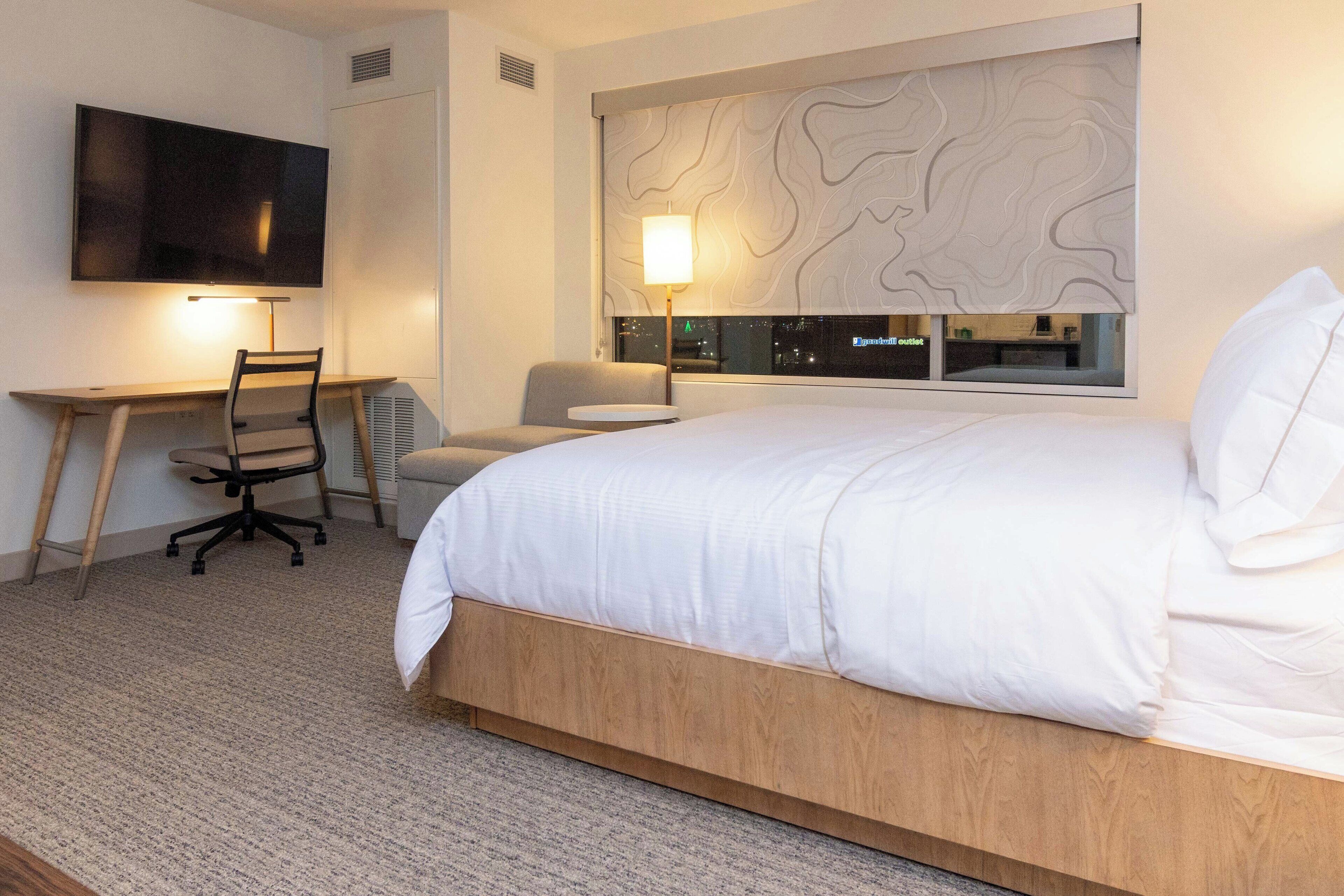 A hotel room with a bed, desk, chair, TV, and a city view through the window.