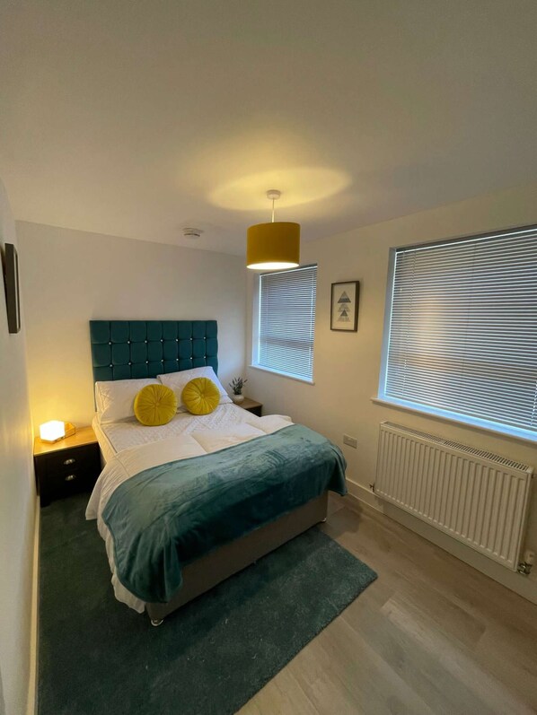 A bedroom with a teal upholstered headboard, a bed with white bedding and two yellow pillows, a bedside table with a lamp, a window with blinds, and a radiator.