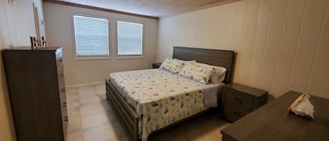 A bedroom with a bed, a dresser, a nightstand, and a desk. There are two windows with blinds, a ceiling light, and a wooden ceiling.