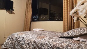 A bedroom with a bed, a window with curtains, a flat-screen TV mounted on the wall, and a vase with flowers on the bed. - Hotel Rincon de la Alameda (Quito)